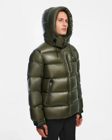 A person wears the Rudsak VINCE S - 6124560 Loden Green men’s down puffer jacket with a hood, zippered pockets, and high collar against a white background. This stylish, weather-resistant jacket is both functional and modern.