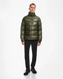 A person stands against a white background wearing the Rudsak VINCE S - 6124560 Loden Green mens down puffer jacket, which has a shiny weather-resistant finish, high collar, front zipper, zippered chest pocket, black pants, and black shoes.