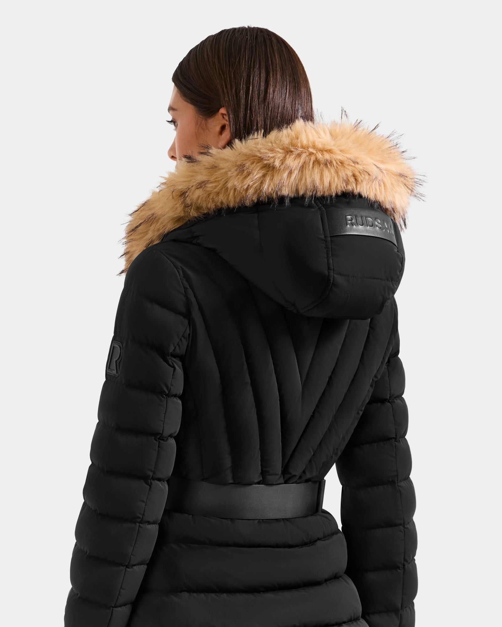 A woman with straight brown hair is seen from behind wearing the Rudsak ALISA F - L125527 black puffer jacket, featuring a wide belt, tan faux-fur trimmed hood, stitched detailing, and a logo patch on the arm.
