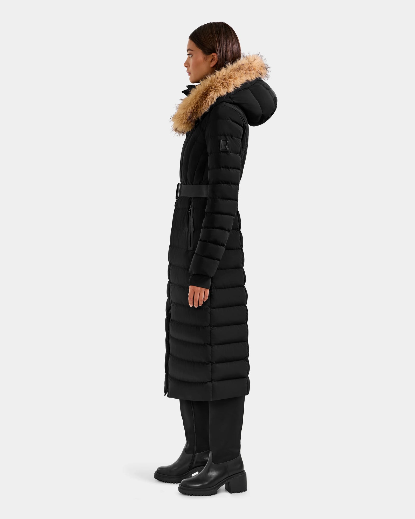 Woman in profile wearing the Rudsak ALISA L - L125574 Black long quilted puffer coat with belted waist and brown faux-fur-trimmed hood, paired with black pants and boots, standing against a plain light background.