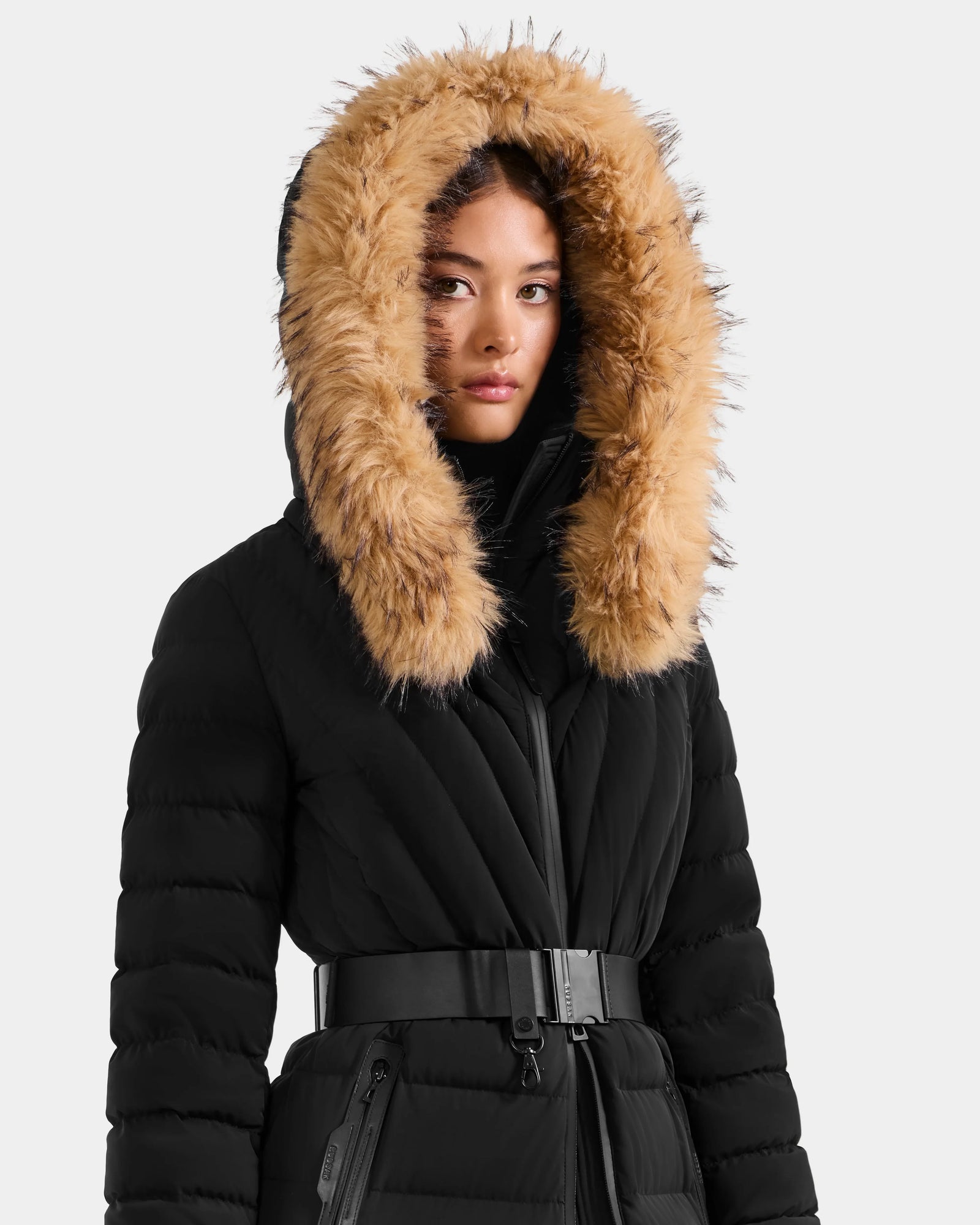 A woman models the Rudsak ALISA L - L125574 Black coat, a black quilted puffer with a belted waist and large hood trimmed with thick tan faux fur, posed against a plain light background.