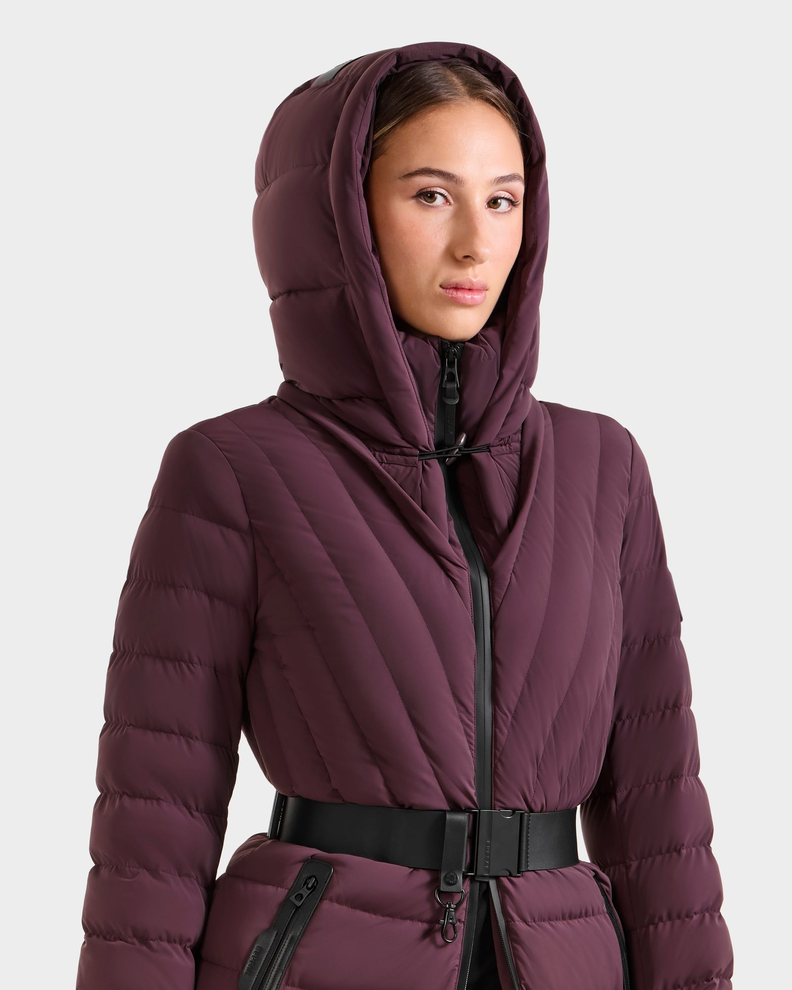 Someone in the Rudsak ALISA M - L125586 Bordeaux parka, with quilted down, a vegan leather belt, and a zipper, stands against a plain light background.