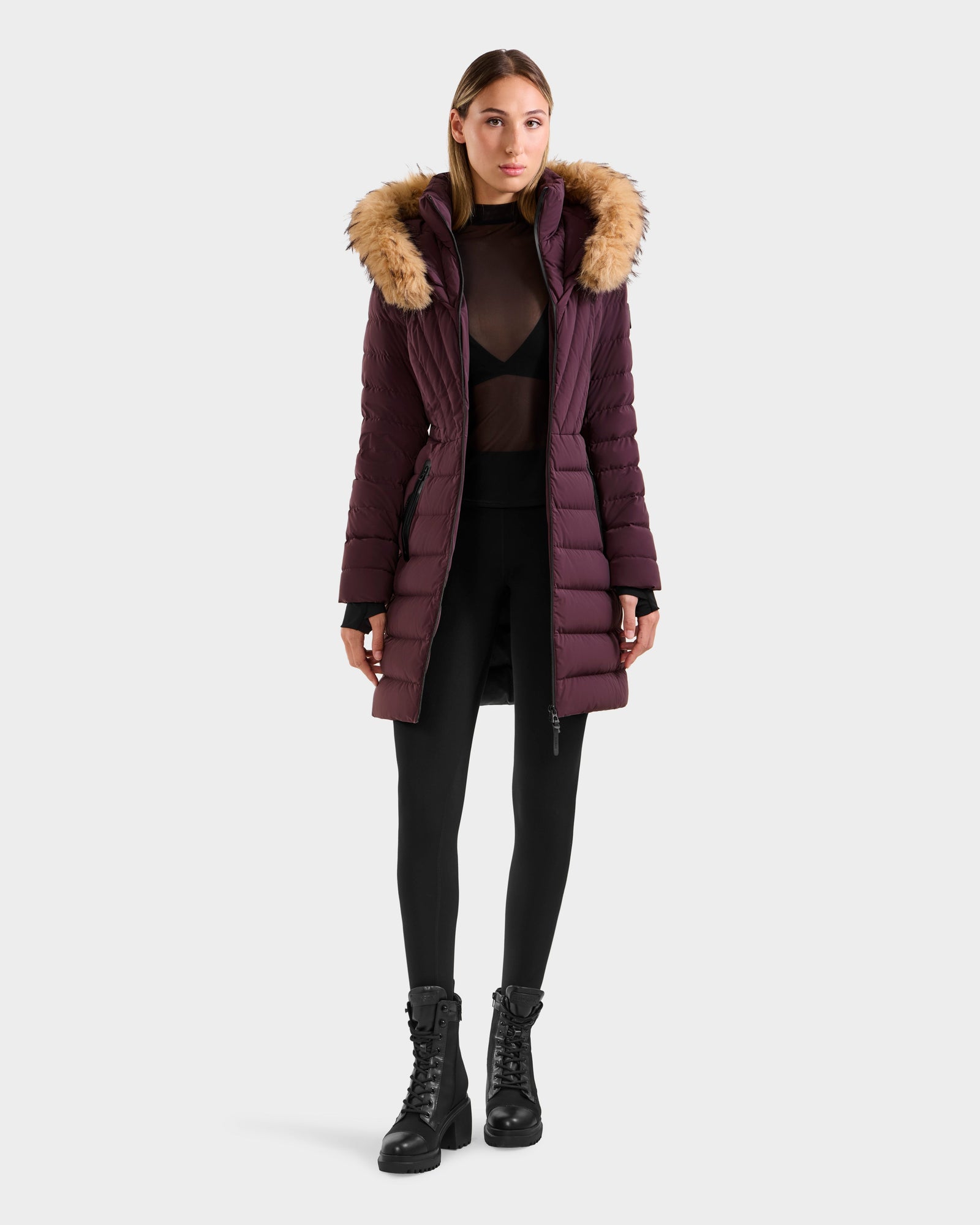 A woman faces forward in a Rudsak ALISA M - L125586 Bordeaux long maroon quilted down parka with faux fur hood, black leggings, sheer top over black bra, and black boots. The background is plain and light-colored.