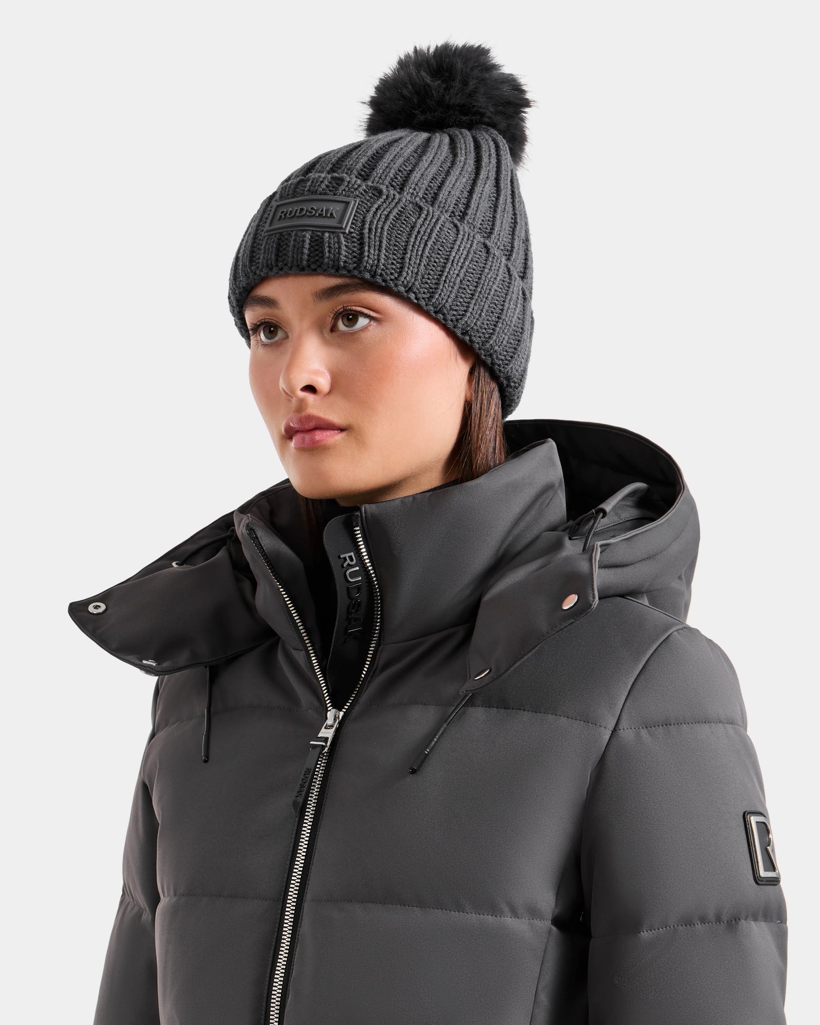 A woman wears the Rudsak ANISA - L423500 Grey quilted jacket with high collar and zipper, paired with a women’s knit pom pom hat, standing against a light background—perfect for a chic winter look.