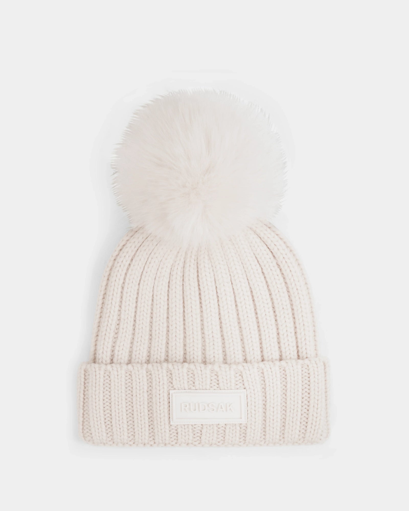 The ANISA - L423500 Vanilla by Rudsak is a cream-colored ribbed knit beanie with a plush faux fur pom pom and folded brim, accented by a rectangular Rudsak label—perfect for elevating your winter wardrobe.