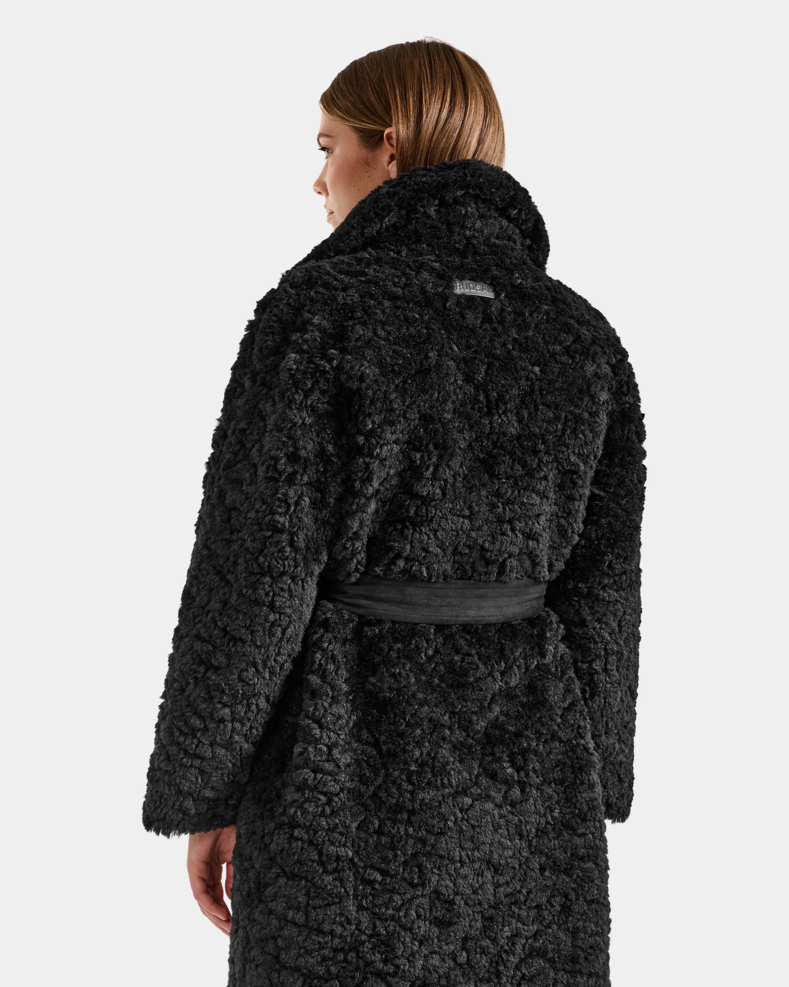 A woman with straight, light brown hair is shown from the back wearing the Rudsak ANNABELLE FSU - L125512 Black women's reversible trench coat with textured faux fur, high collar, and black belt against a plain light background.