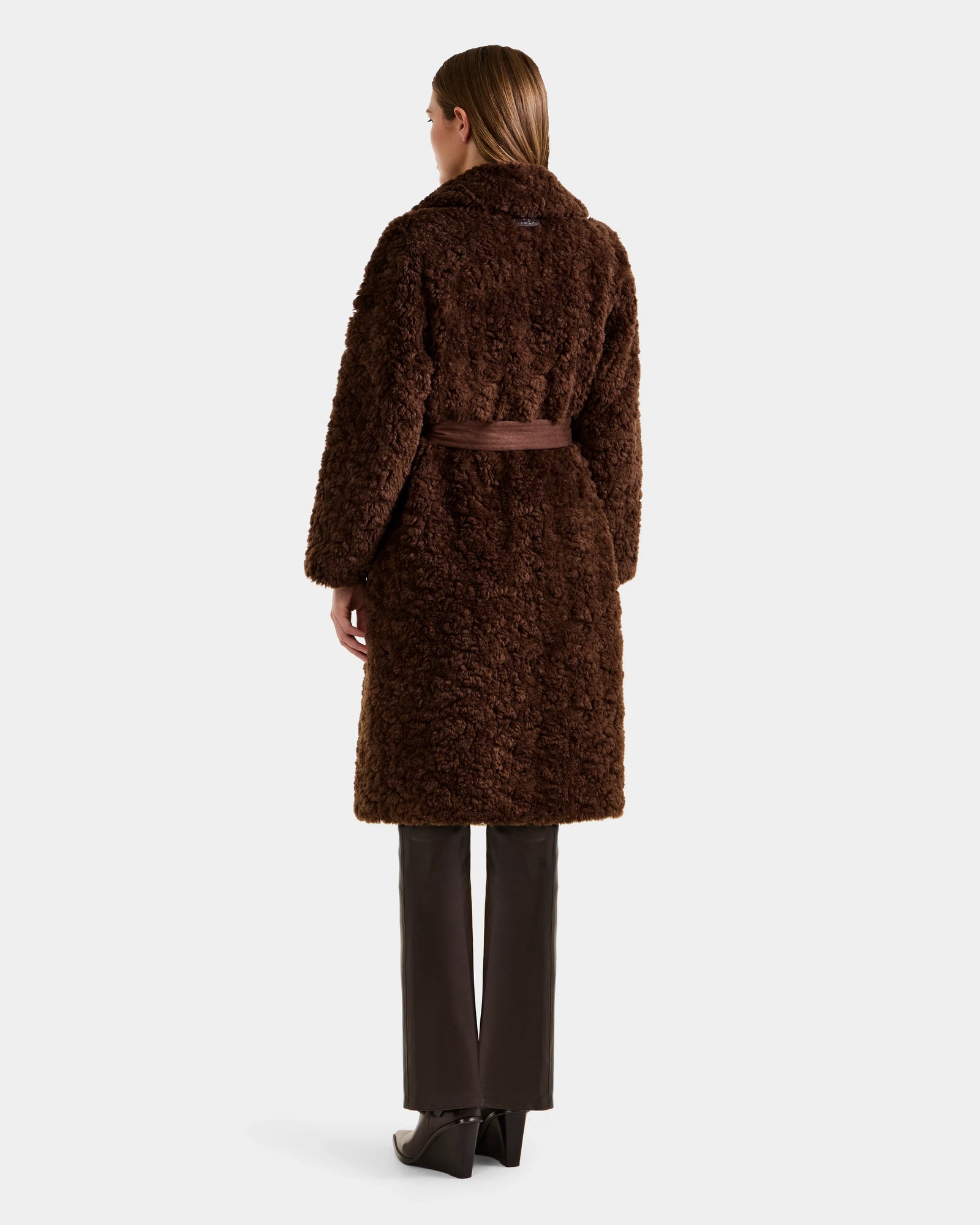 A person with straight brown hair models the Rudsak ANNABELLE FSU - L125512 Espresso sherpa teddy coat, knee-length with a belt, paired with dark pants and black heeled shoes, shown from the back against a plain background.