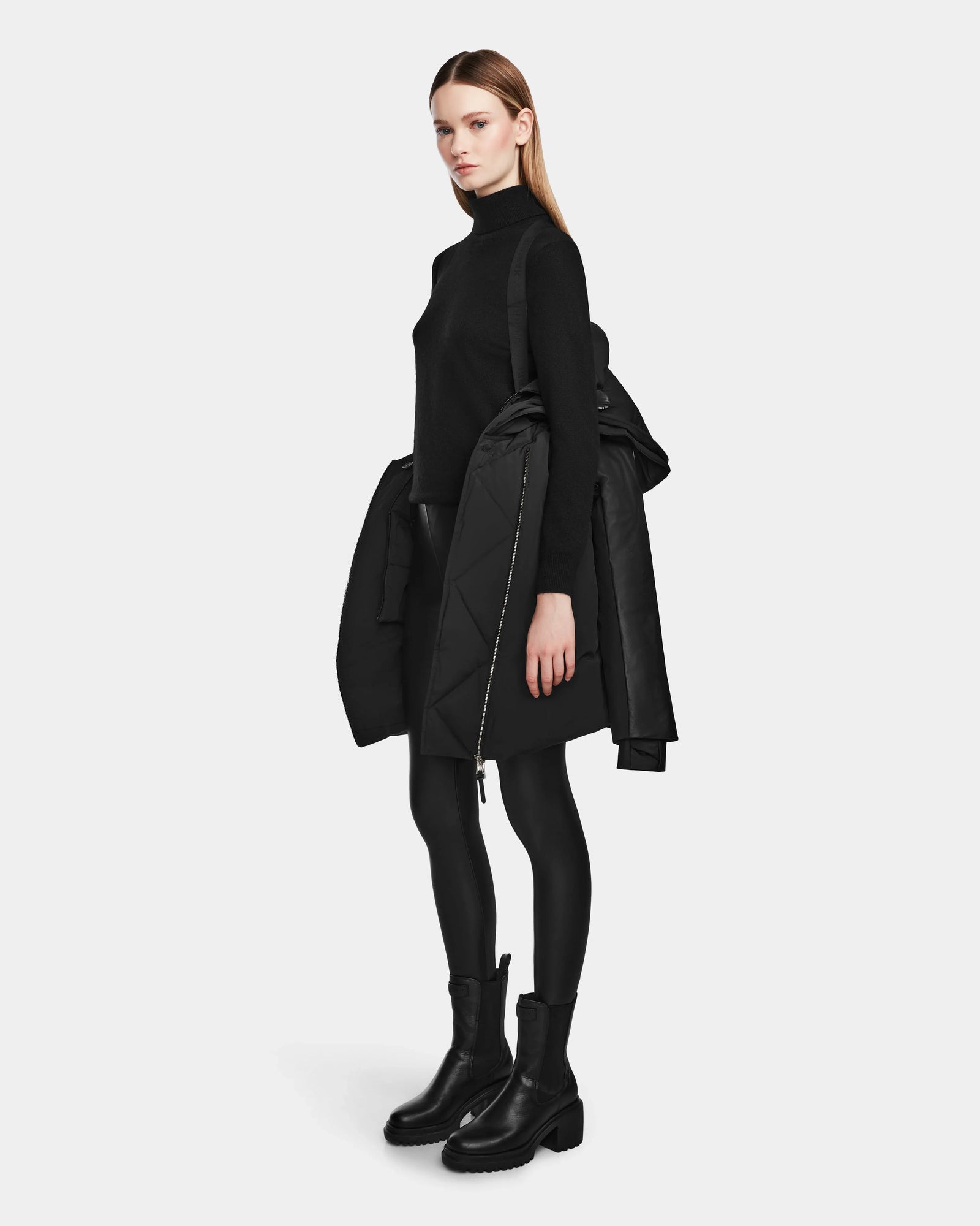 A woman stands sideways against a white background, holding the Rudsak AURELLE - L124538 Black women’s down parka with vegan leather sleeves over her arm while wearing a black turtleneck, skirt, leggings, and boots.