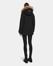 A person wearing the Rudsak AURORE - L123561 Black/Natural coat stands facing away, showcasing its fitted black design with fur trim against a plain light background.