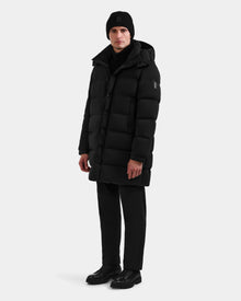 A person showcases winter versatility wearing the Rudsak BRAYDEN M-M - M125535 Black men’s down puffer coat, styled with black pants, a black knit beanie, and black shoes against a plain white background.