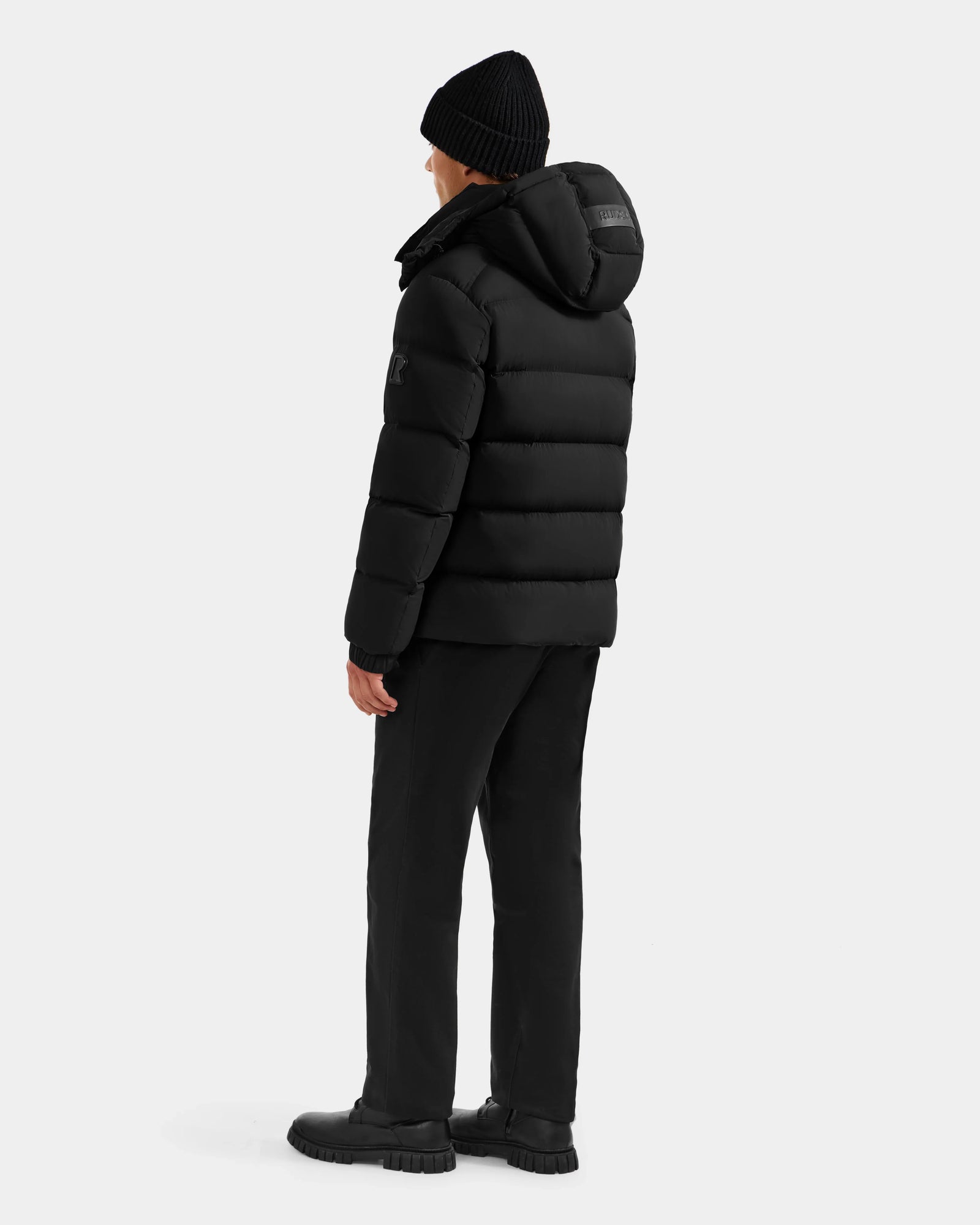 A person stands facing away against a plain light background, wearing the Rudsak BRAYDEN M-S - M125524 Black RDS-certified down winter jacket with hood, black pants, boots, and a black knit beanie.