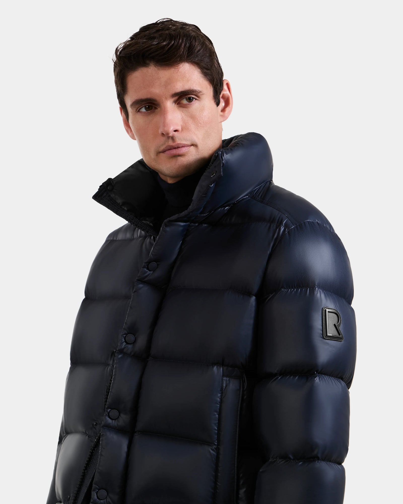 A man stands against a plain light background wearing the Rudsak BRAYDEN S - M125525 Navy men's quilted down puffer jacket, featuring a shiny dark blue finish, high collar, and black R patch on the sleeve.