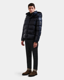 A man faces slightly to the side, wearing the Rudsak BRAYDEN S - M125525 Navy men's quilted down puffer jacket with hood, dark cuffed pants, and brown suede boots against a plain white background.