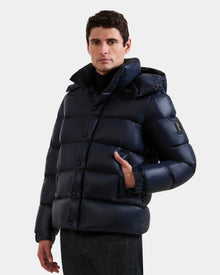 A man faces forward with one hand in his pocket, wearing the Rudsak BRAYDEN S - M125525 Navy quilted down puffer jacket with a hood and black pants against a plain white background.