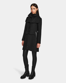 A woman with long brown hair and a neutral expression wears the Rudsak CAROLINE - L124543 Black, a knee-length premium wool-blend coat with a cape-like collar, paired with black leggings and boots against a plain background.
