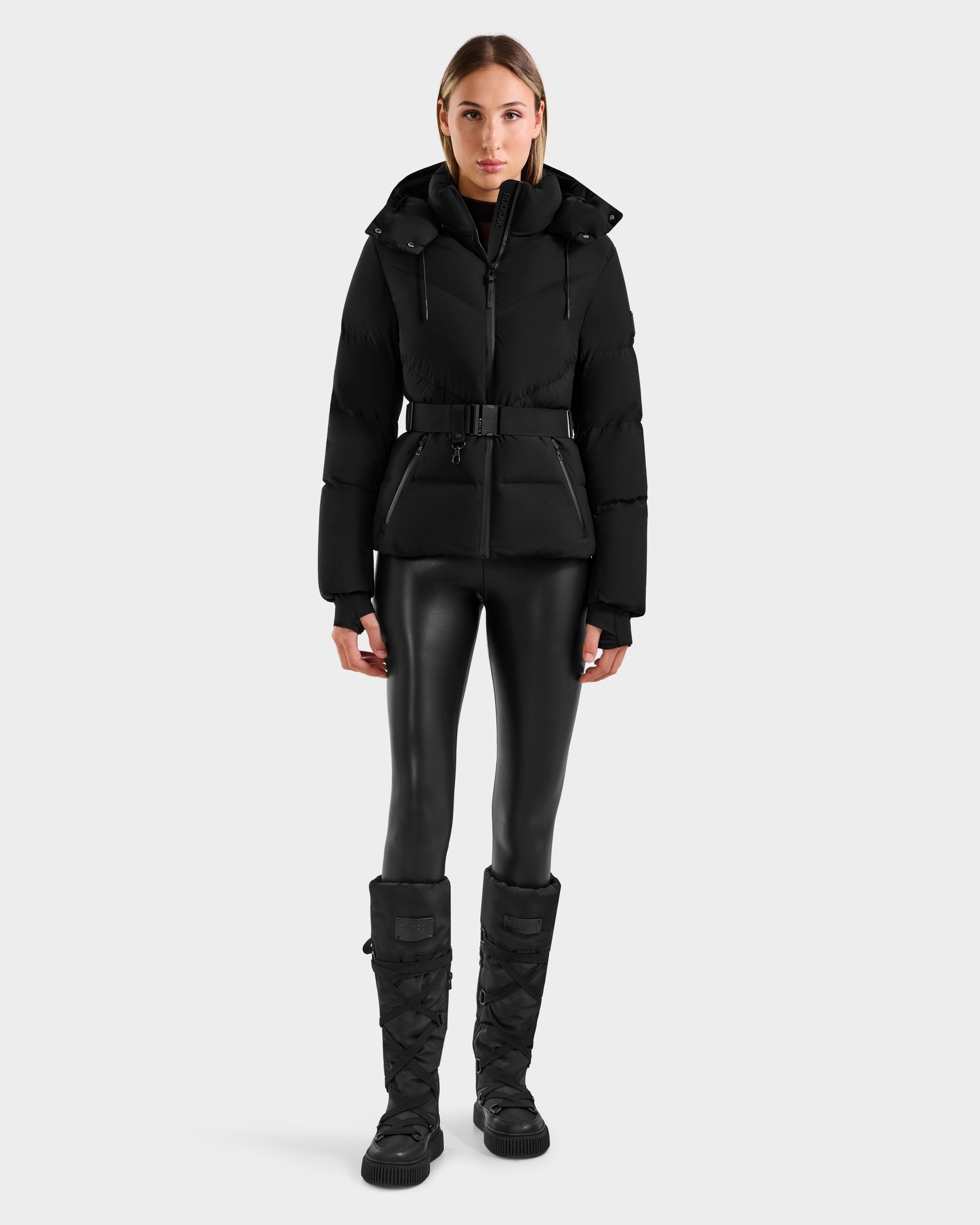 A woman faces forward wearing the Rudsak CHRISTELLE M - L125542 Black quilted down puffer jacket with belt, paired with shiny black leggings and knee-high winter boots against a plain light gray background.