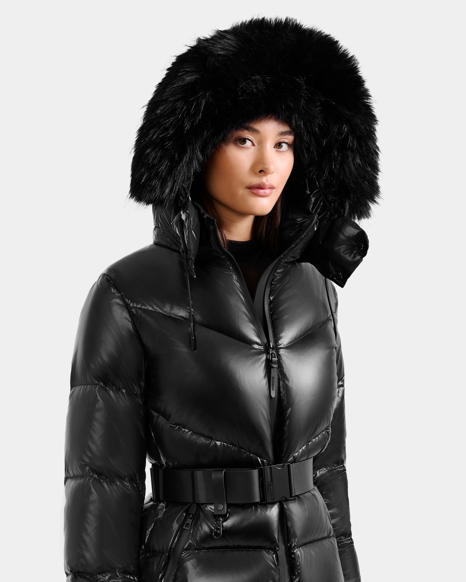 A woman wears the Rudsak CHRISTELLE S - L125544 Black down puffer with a large faux fur trim hood and black belt, standing against a plain light background.