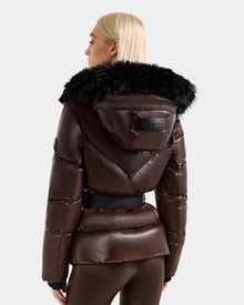 A person with blond hair wears the Rudsak CHRISTELLE S - L125544 Espresso women’s down puffer jacket featuring a black faux fur hood trim, black belt, and a RUDSAK patch on the back. The background is plain and light-colored.