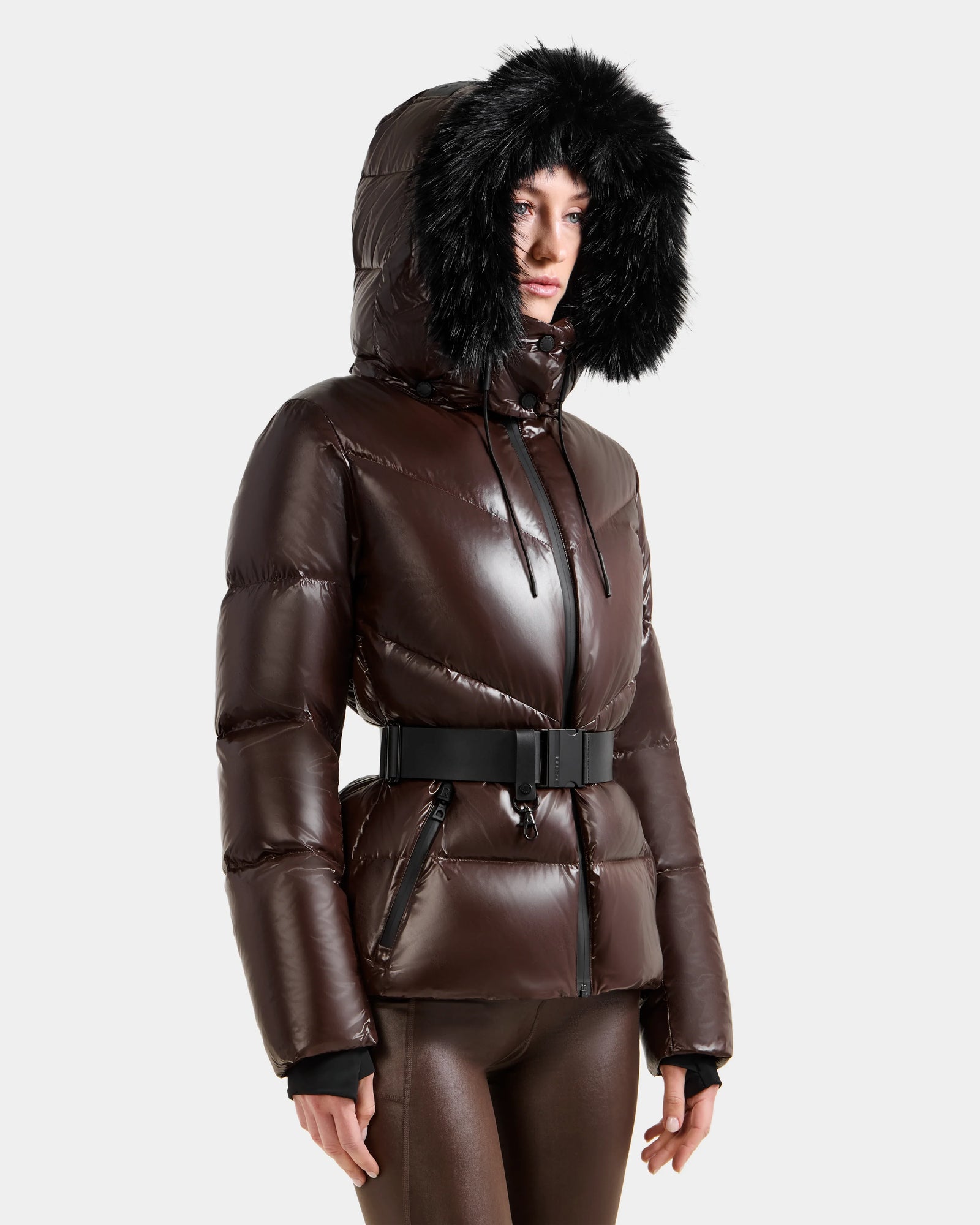 A person wears the Rudsak CHRISTELLE S - L125544 Espresso, a shiny brown women’s down puffer with black faux fur trim hood and black belt, paired with matching brown pants against a plain light background.