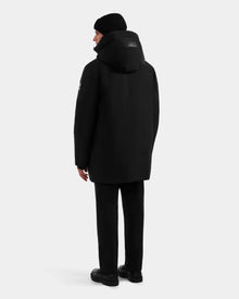 A person wearing the Rudsak COREY - M125512 Black men’s down parka, paired with black pants, shoes, and a beanie, stands facing away from the camera against a plain light gray background.