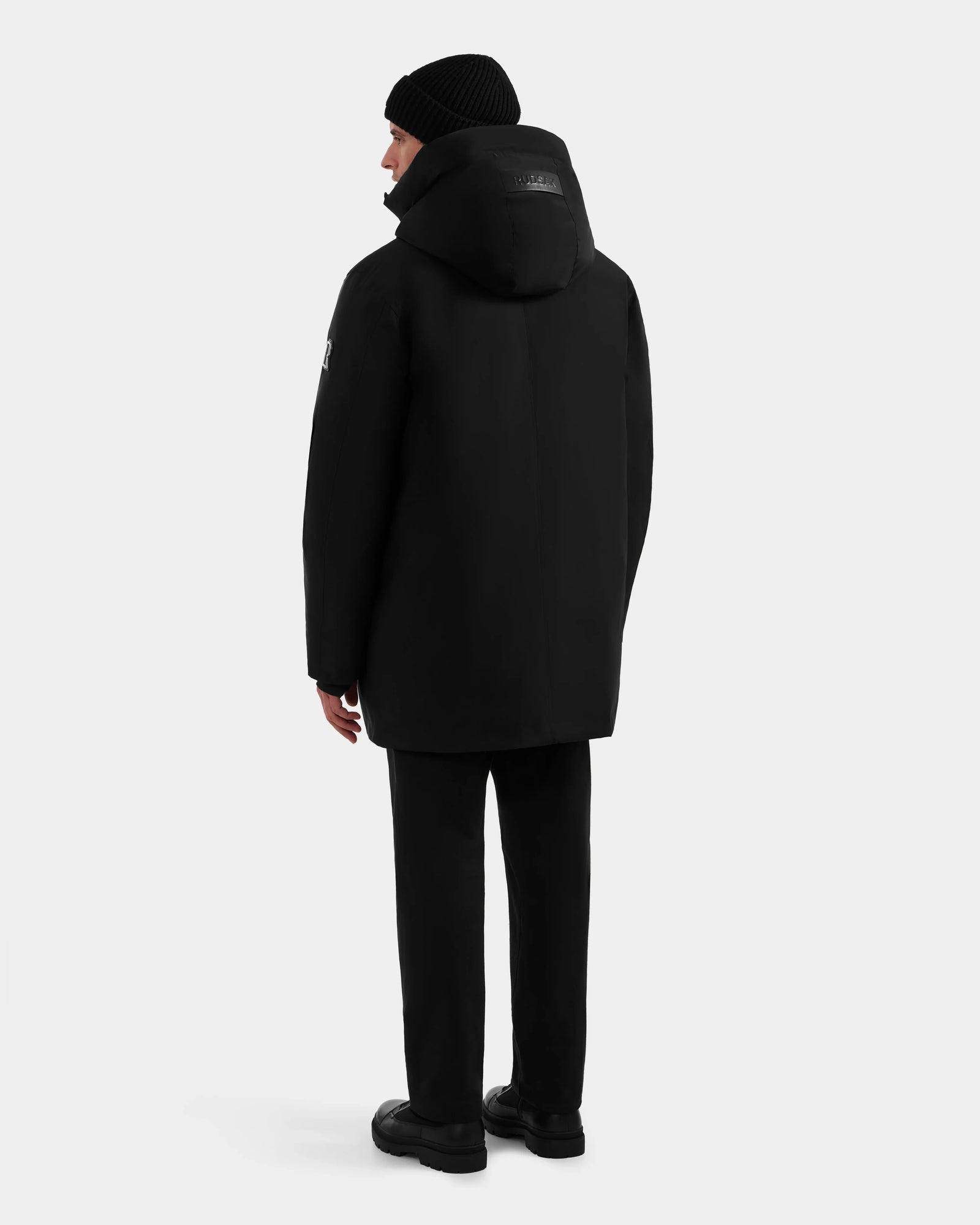 A person wearing the Rudsak COREY - M125512 Black men’s down parka, paired with black pants, shoes, and a beanie, stands facing away from the camera against a plain light gray background.
