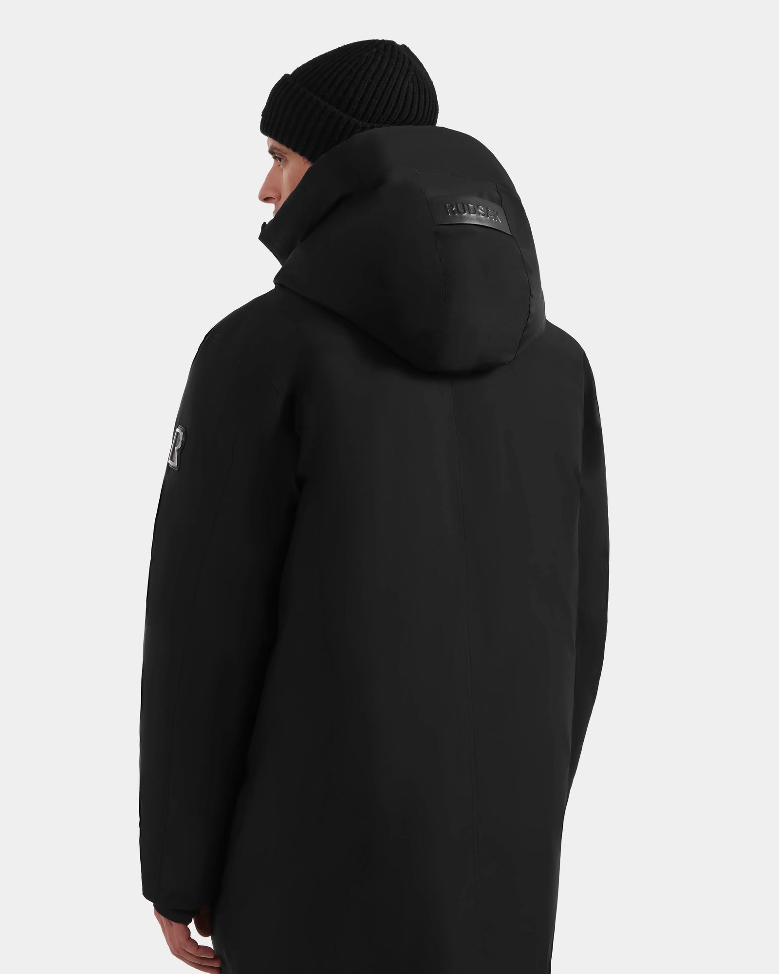 A person is shown from the back and slightly to the side, wearing the Rudsak COREY - M125512 Black men’s down parka with a black knit beanie, against a plain light gray background.