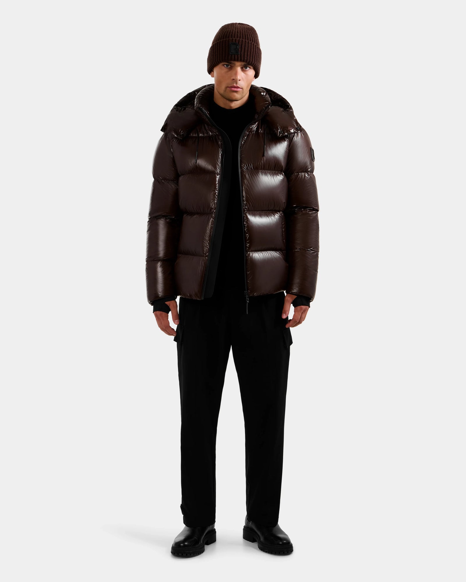 A person wearing the Rudsak DANIEL S - M124508 Espresso men's hooded down puffer jacket with a glossy, weather-resistant shell stands against a plain background, paired with a brown knit beanie, black pants, and black shoes. Arms rest at their sides.