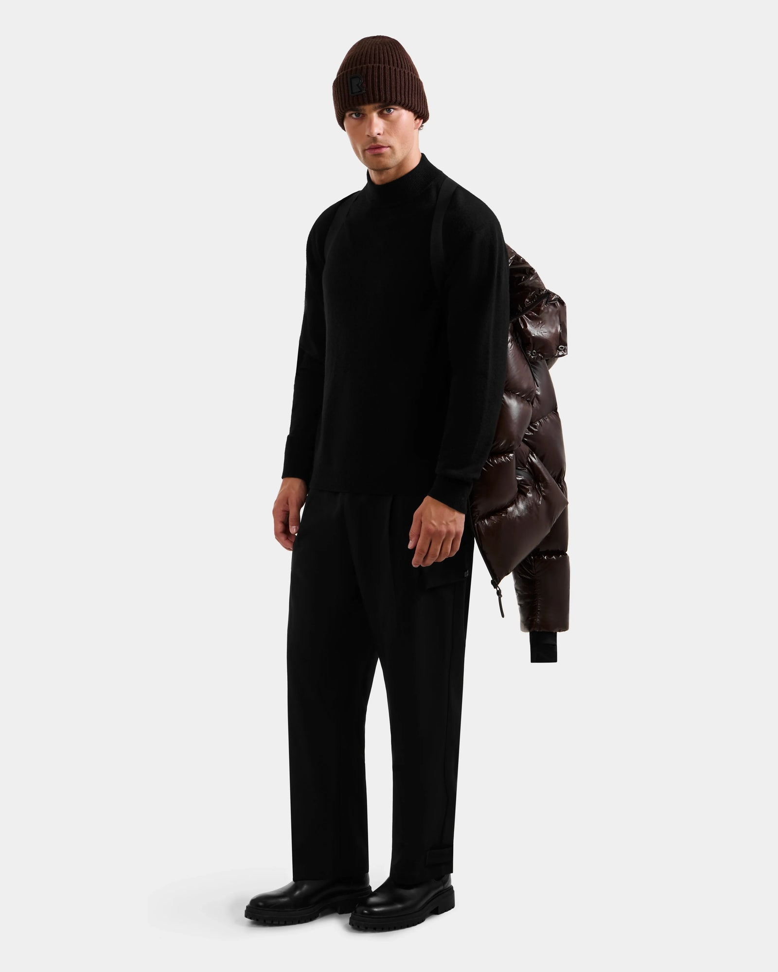 A man in a black turtleneck, wide-leg pants, and brown knit beanie stands against a white background with the Rudsak DANIEL S - M124508 Espresso weather-resistant puffer draped over his shoulder.
