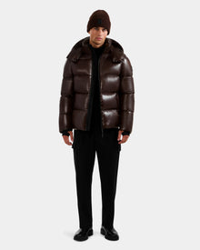 A person faces forward in a Rudsak DANIEL S - M124508 Espresso glossy, weather-resistant puffer jacket with high-loft down fill, paired with black pants, black shoes, and a brown knit beanie against a plain light background.