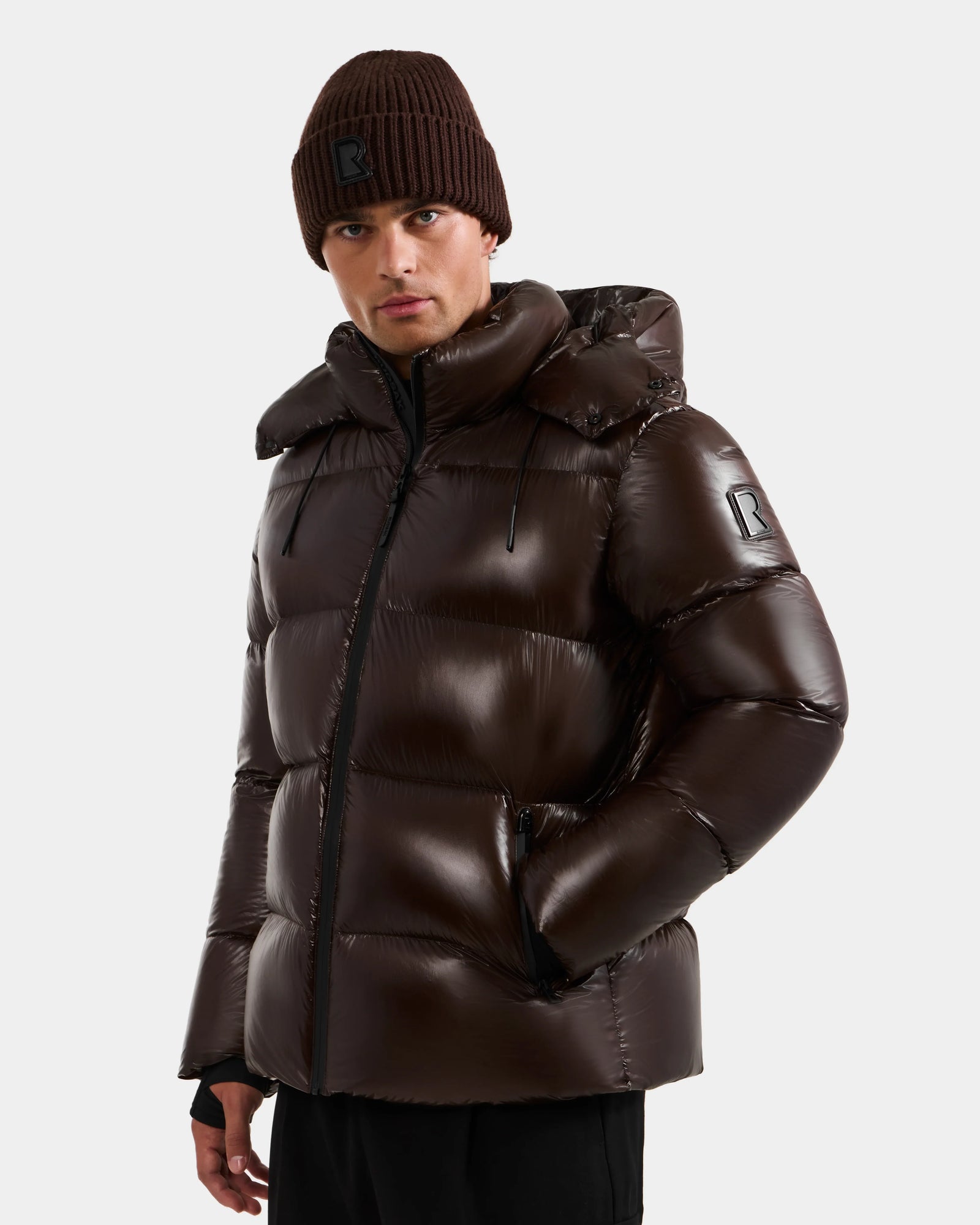 A man wears the Rudsak DANIEL S - M124508 Espresso puffer, featuring a weather-resistant glossy shell and stitched letter R logo, paired with a matching brown knit beanie, both against a plain light background.