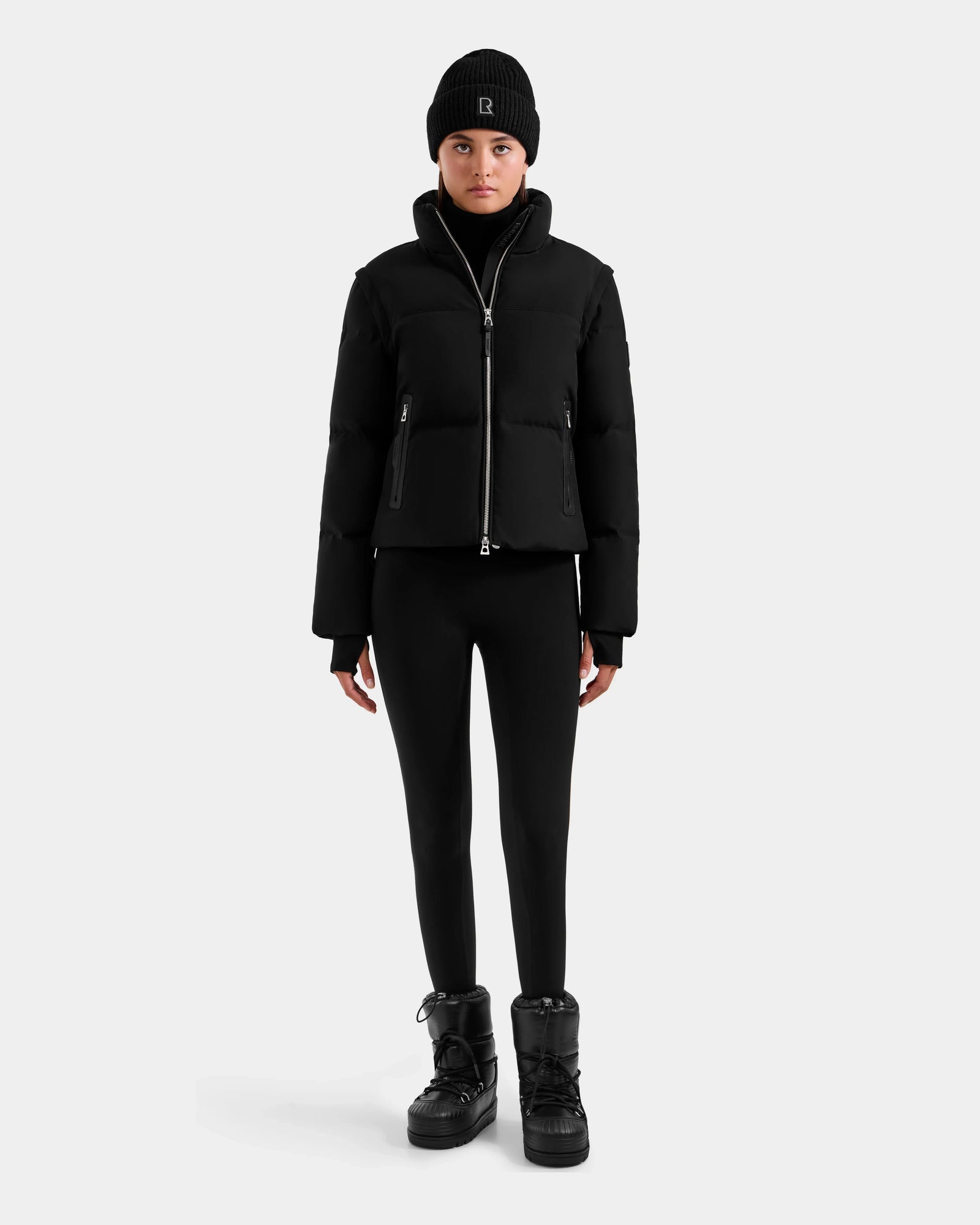 A person faces forward against a light gray background, wearing the Rudsak DELILAH M - L125538 Black women's down puffer with removable sleeves, black leggings, and black winter boots.