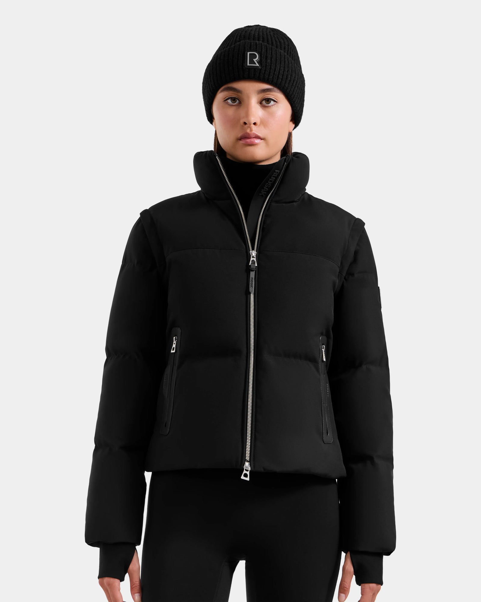 A person wearing a black Rudsak DELILAH M - L125538 women’s down puffer jacket, a black beanie with a small white logo, and black leggings stands facing forward against a plain light background.