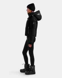 A woman in profile wears the Rudsak DELILAH M - L125538 Black down puffer with removable sleeves, paired with black leggings, winter boots, and a black knit beanie, standing against a plain light background.
