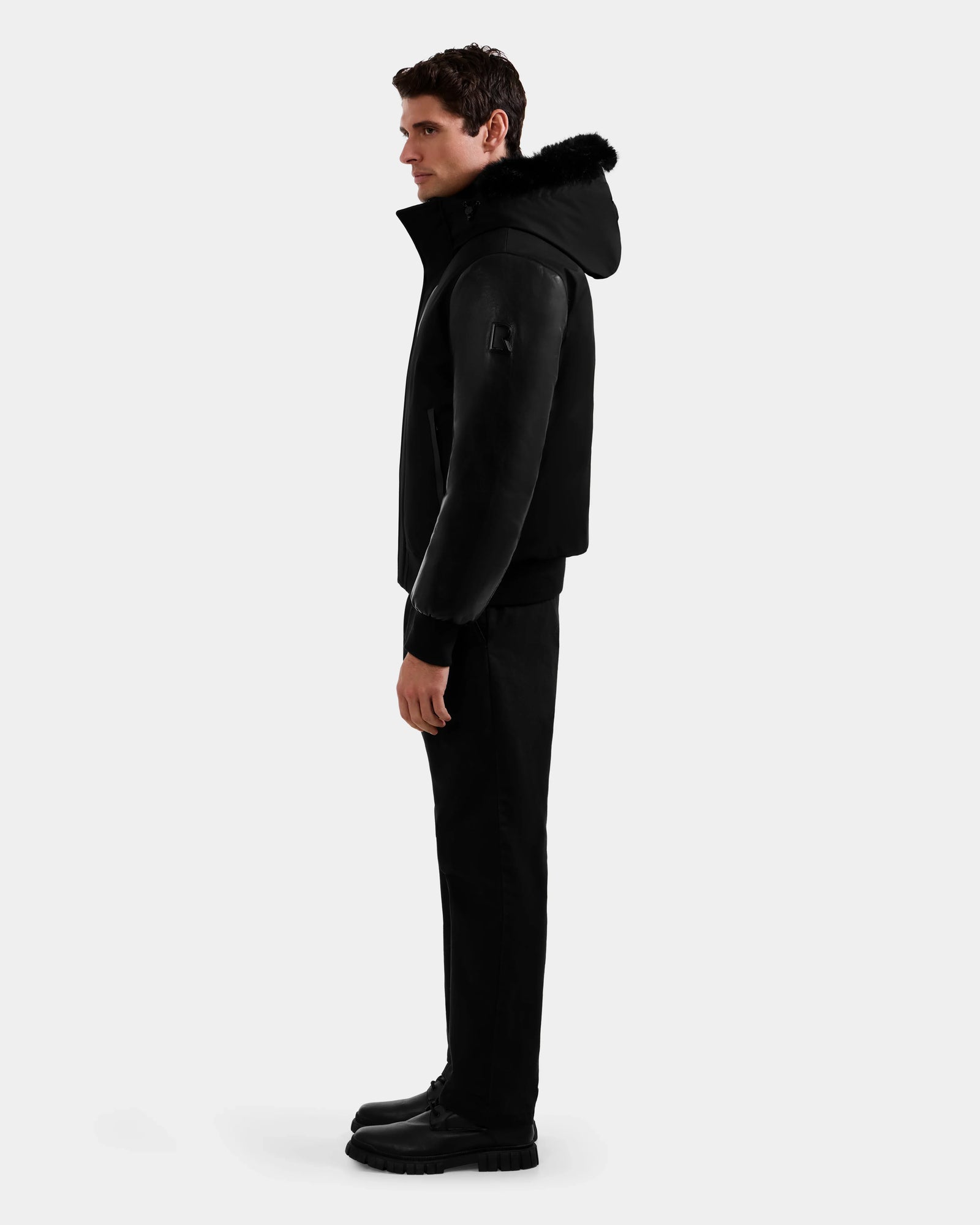 A man in profile stands against a plain background, wearing the Rudsak DERON - M125539 Black men’s down bomber with fur trim and vegan-leather sleeves, paired with black pants and boots.