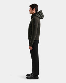 A man stands in profile against a plain background, wearing the Rudsak DURIN M - M125530 dark olive men’s hooded jacket with lightweight down insulation, paired with black pants and boots. He faces left with his arms relaxed at his sides.