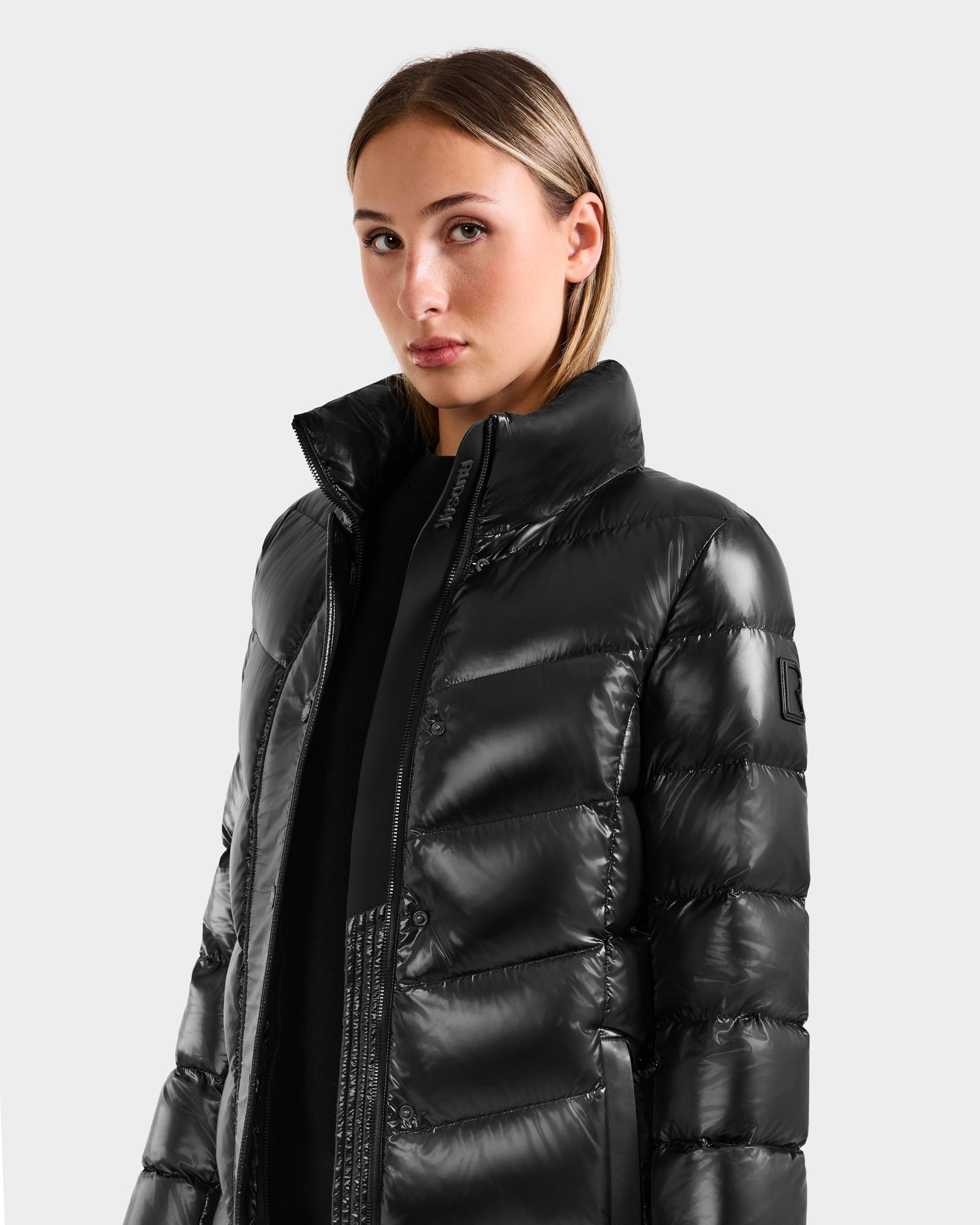 A woman with straight, light brown hair wears the Rudsak ELISA S - L125572 Black shiny quilted puffer jacket with a high collar, standing against a plain light gray background.