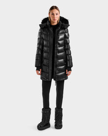 A woman faces forward against a plain light gray background, wearing the Rudsak ELISA S - L125572 Black shiny mid-length winter coat with a fur-trimmed hood, paired with black leggings and black winter boots.