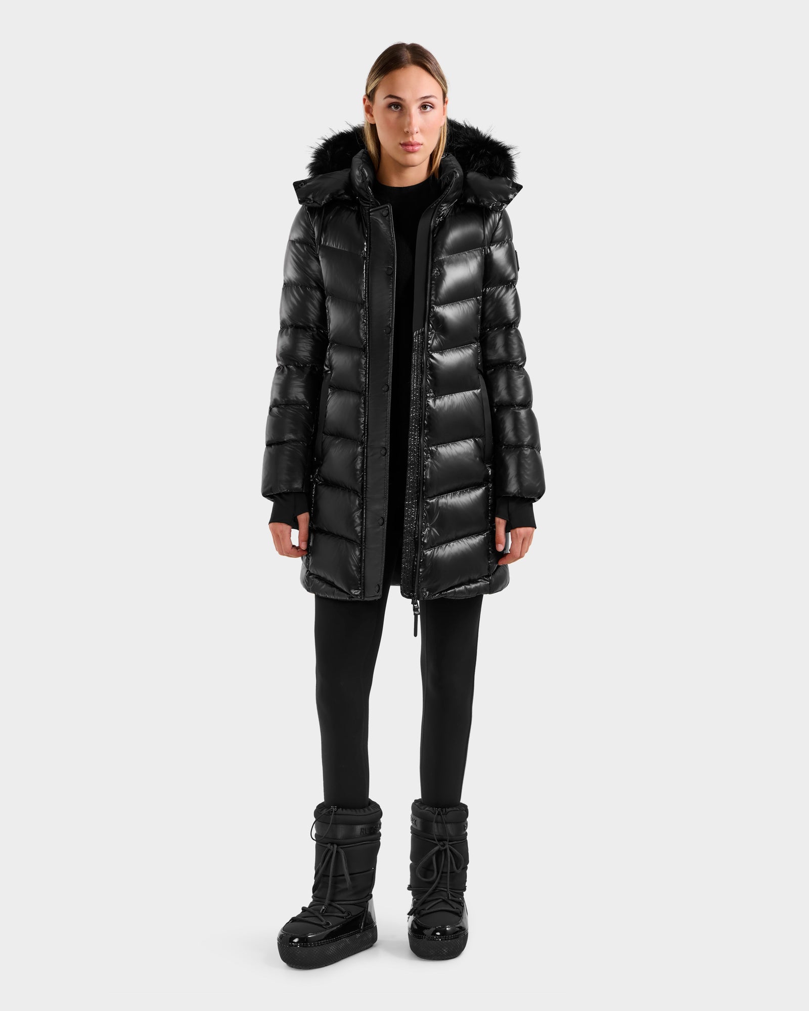 A woman faces forward against a plain light gray background, wearing the Rudsak ELISA S - L125572 Black shiny mid-length winter coat with a fur-trimmed hood, paired with black leggings and black winter boots.