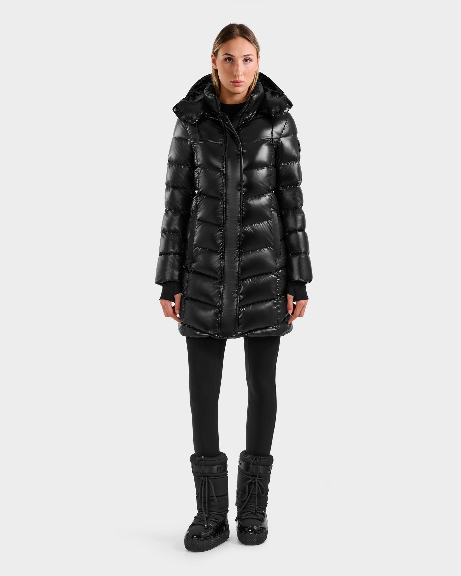A person faces forward wearing the Rudsak ELISA S - L125572 Black shiny quilted puffer jacket with hood, paired with black leggings and winter boots, against a plain light gray background.