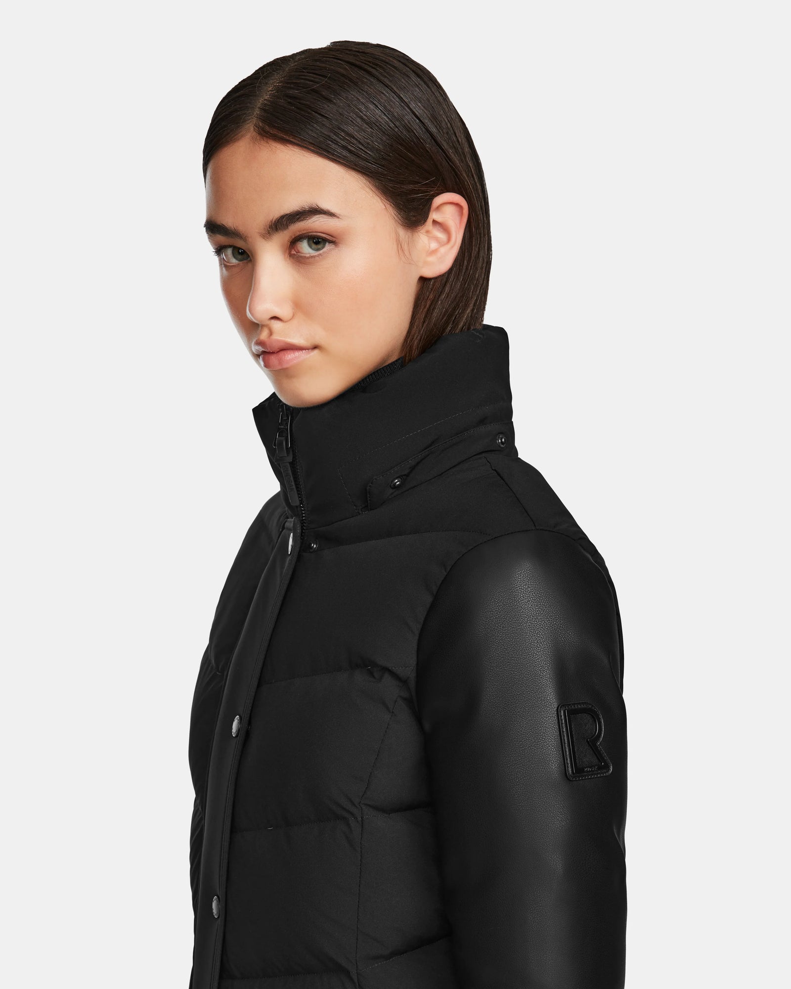 A person with straight dark hair wears the Rudsak ELSA - L123535 Black, a women’s quilted down puffer with leather sleeves and a B patch on the high collar, looking over their shoulder against a plain light background.