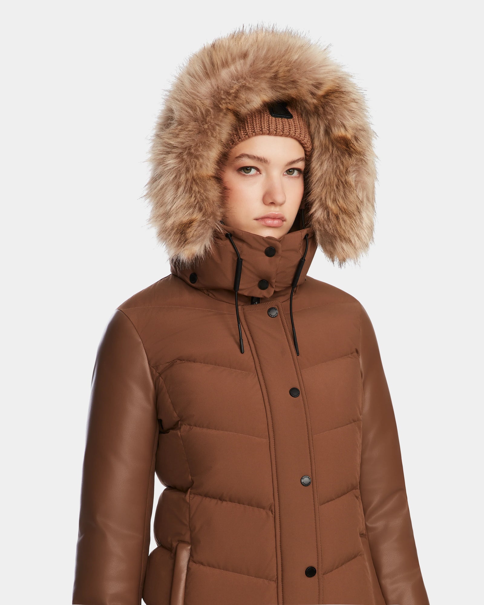 A woman in the Rudsak ELSA - L123535 Taupe quilted down puffer with leather sleeves, a high collar, and faux fur-trimmed hood pairs it with a matching knit hat against a light background—highlighting its sustainable design.