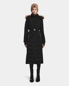 A woman stands facing forward against a plain white background, wearing the Rudsak ELSIE M - L123517 Black/Natural maxi down puffer with belt and fur-trimmed hood, paired with black boots and a tan knit beanie.