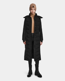 A person wearing the Rudsak ELSIE M - L123517 Black/Natural maxi down puffer, a tan sweater, black pants, black boots, and a tan beanie stands facing forward against a plain white background.