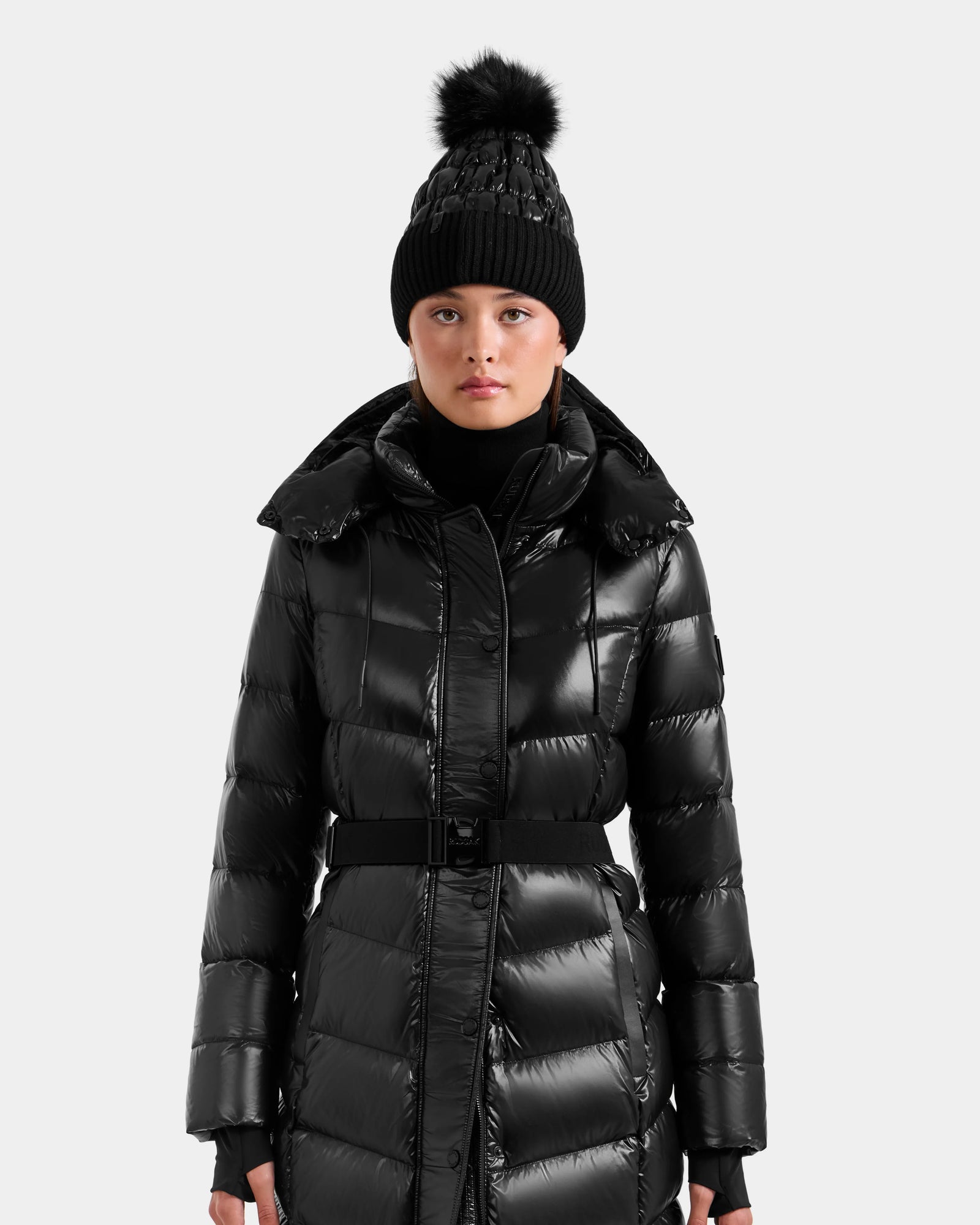 A person models the Rudsak ELSIE S - L124536 Black women’s maxi down puffer coat with a belt and a black knit beanie with a large pom-pom, standing against a plain light gray background.