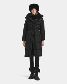 A person stands facing forward wearing the Rudsak EMBER M-L - L125543 Black women’s long down puffer coat with a removable hood, paired with black pants, lace-up fur-trimmed boots, a furry hat, and a scarf against a plain white background.