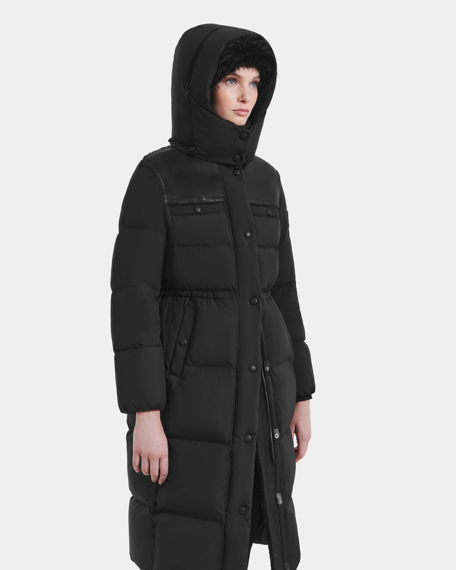 A person wearing the Rudsak EMBER M-L - L125543 Black women's long down puffer coat with a removable hood, visible buttons, and two chest pockets stands slightly to the side against a plain white background.