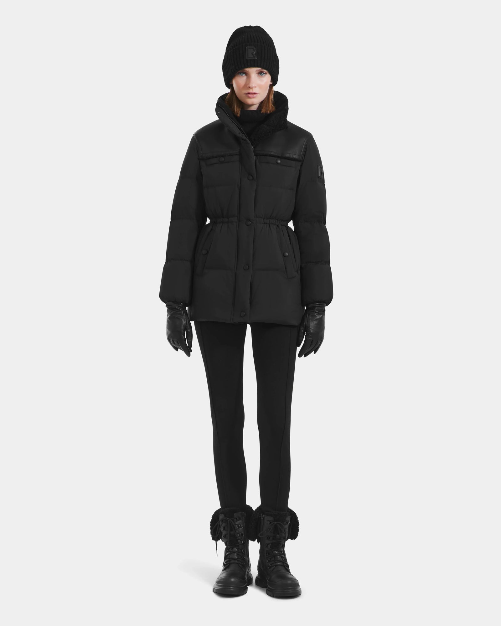 A person stands facing forward against a plain white background, wearing the Rudsak EMBER M-M - L125524 Black women's down puffer with vegan-leather piping, paired with a beanie, gloves, leggings, and lace-up boots.