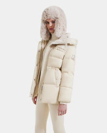 A woman wearing the Rudsak EMBER M-M - L125524 Vanilla down puffer jacket, a matching faux fur ear flap hat, and cream pants stands against a plain white background.
