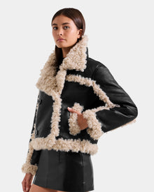 A woman models the Rudsak EMERY - L125548 Black vegan-leather jacket with cream faux shearling trim and a matching black mini skirt, standing against a plain light background.
