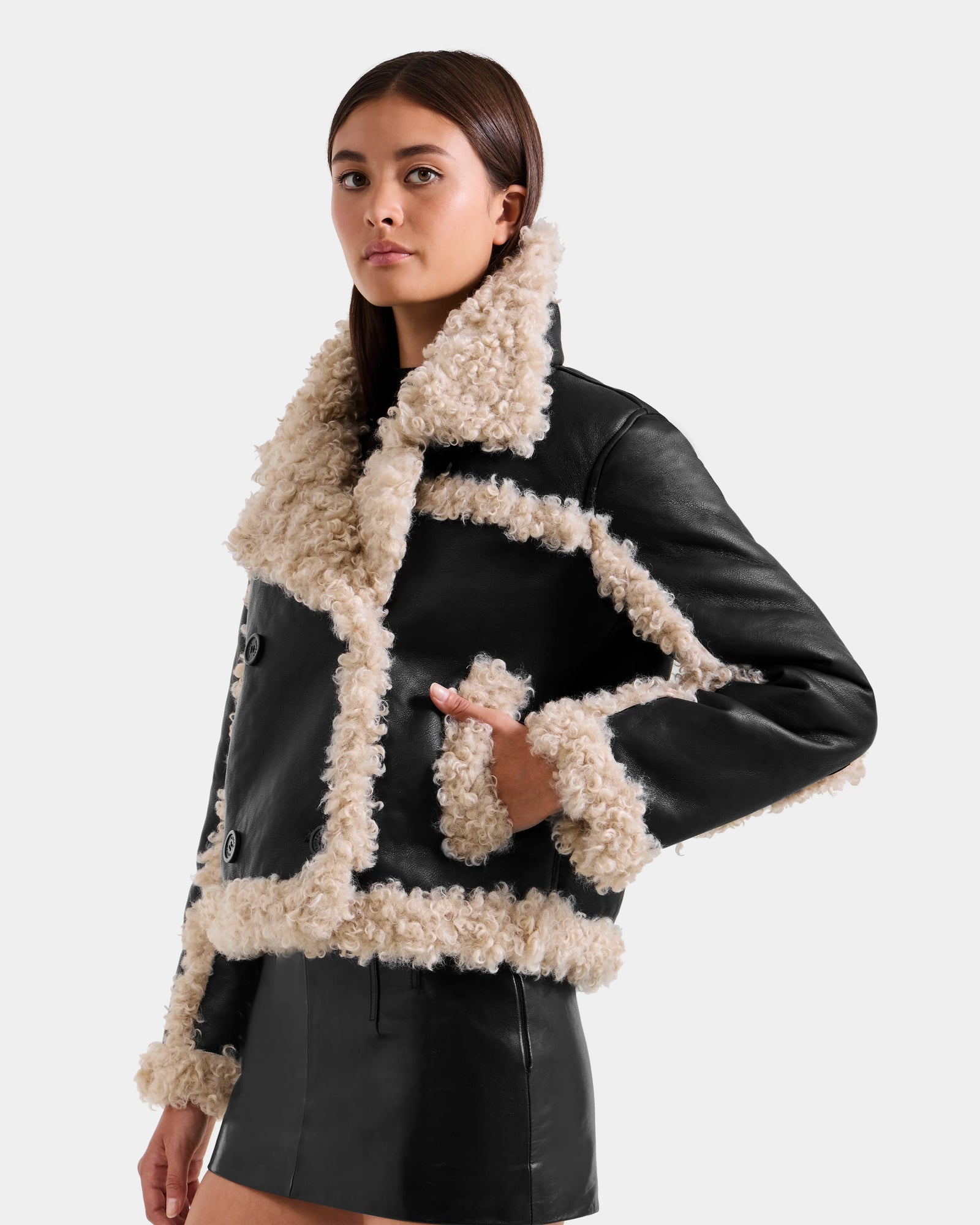A woman models the Rudsak EMERY - L125548 Black vegan-leather jacket with cream faux shearling trim and a matching black mini skirt, standing against a plain light background.